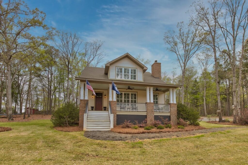 Two-story brick house with front porch, white columns, and American flag on wooded lot.
