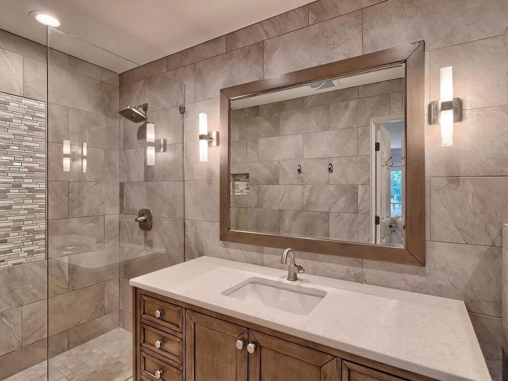 Modern bathroom with marble tile walls, wooden vanity, framed mirror, glass shower enclosure, and recessed lighting.