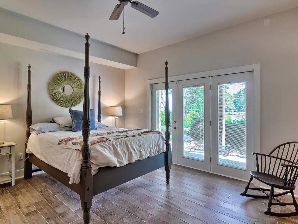 Master bedroom with four-poster bed, ceiling fan, decorative circular wall art, bedside lamps, glass doors, and wooden rocking chair.