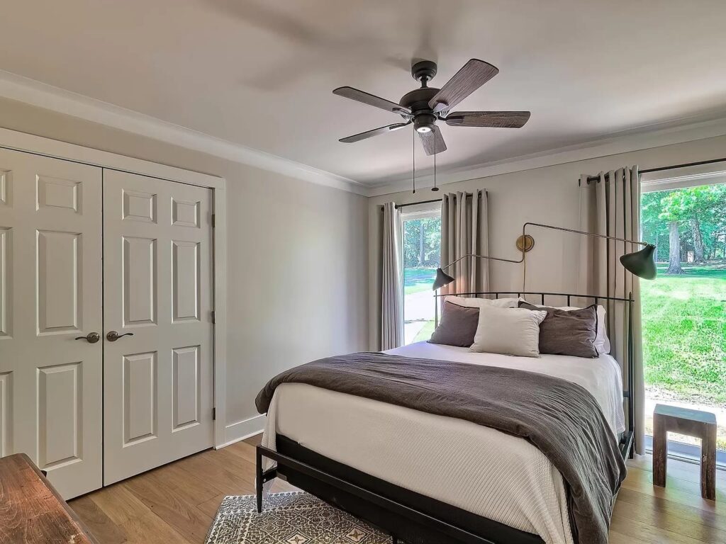 Modern bedroom with black metal bed frame, ceiling fan, white closet doors, and large windows overlooking green lawn.