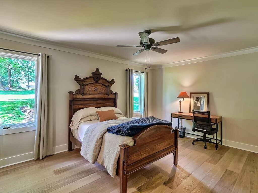 Bright bedroom with ornate wooden bed, ceiling fan, desk, and large windows overlooking green lawn.