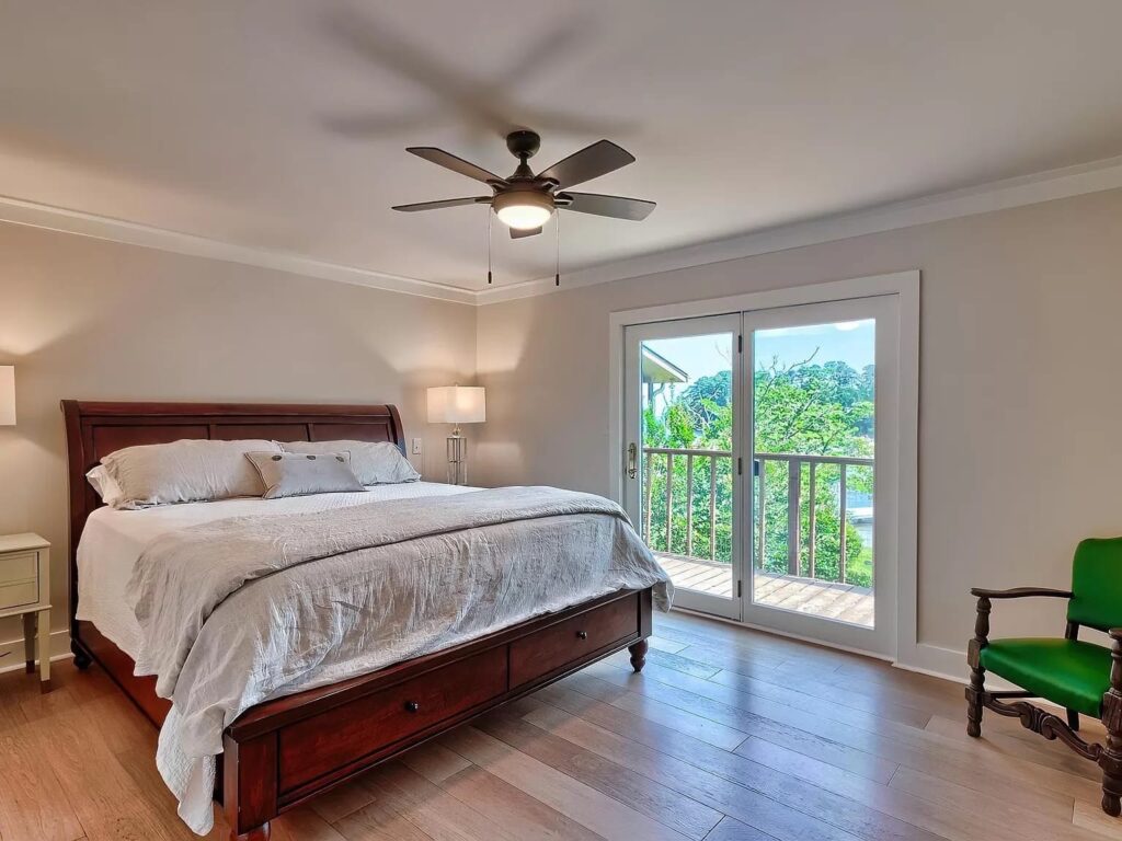 Spacious bedroom with wooden bed, ceiling fan, hardwood floors, and glass doors opening to a balcony overlooking water and trees.
