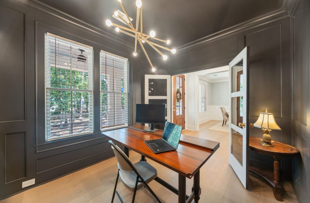 Modern home office with dark gray walls, wooden desk, brass chandelier, and large windows overlooking greenery.