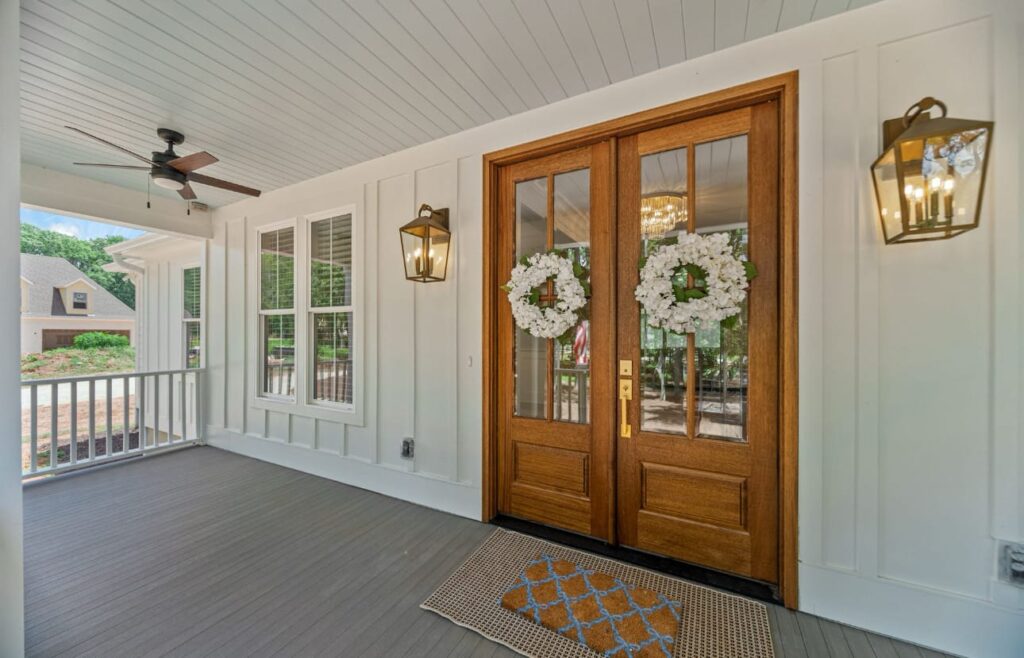 Elegant covered porch with wooden double doors, white windows, ceiling fan, and floral wreaths on the entrance.