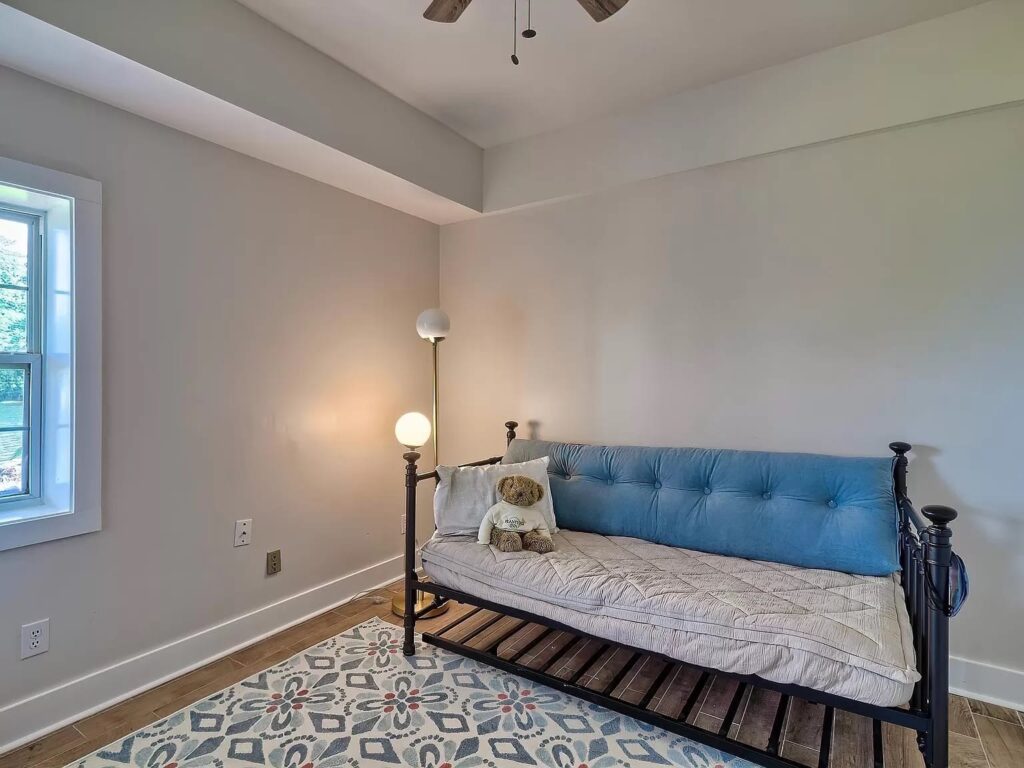 A modern bedroom with a black metal daybed, blue upholstered headboard, and white mattress, decorated with a teddy bear and pillows, featuring hardwood floors and a pendant light.
