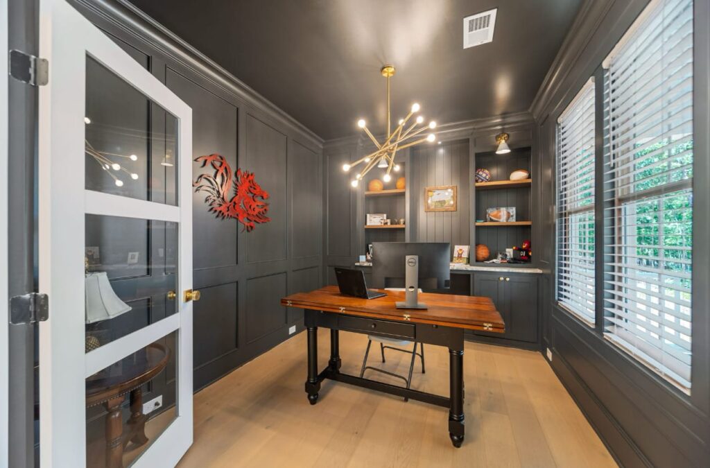 Modern home office with dark gray walls, wooden desk, built-in shelving, and gold pendant lighting.