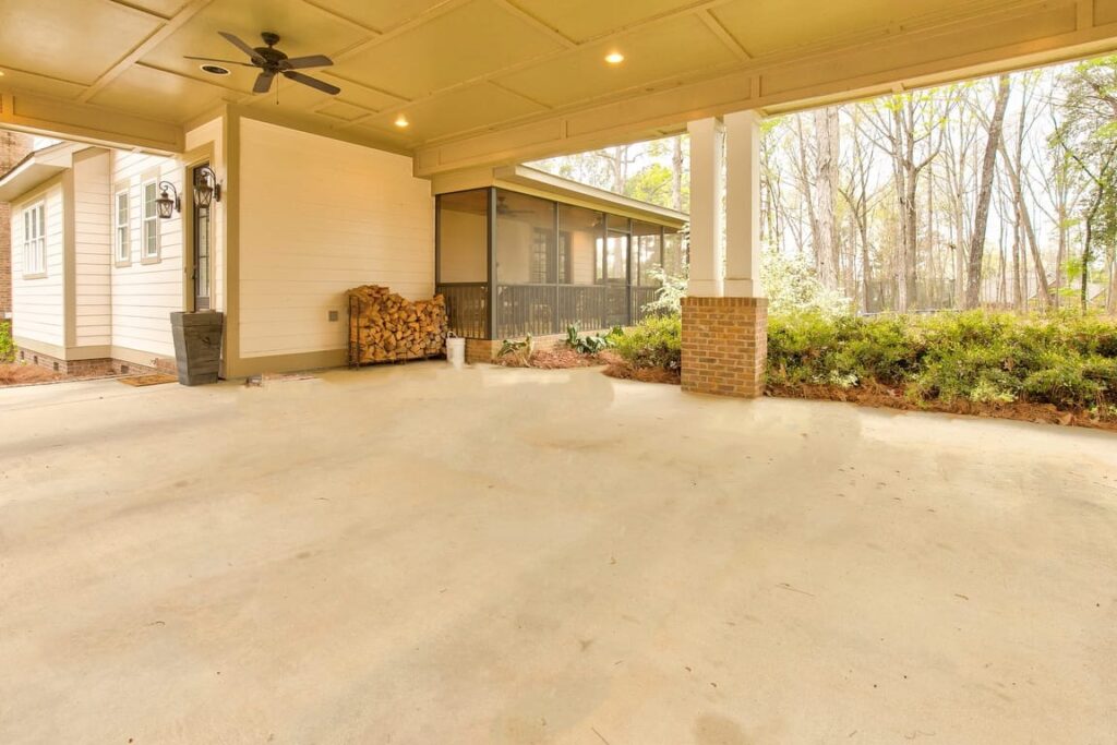 Spacious covered patio with concrete floor, ceiling fan, and brick columns overlooking wooded landscape.