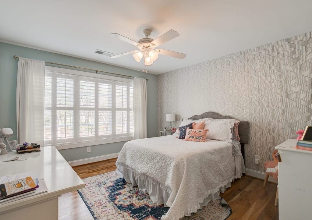 Bright bedroom with bed, ceiling fan, white desk, shuttered windows, and geometric patterned wallpaper.