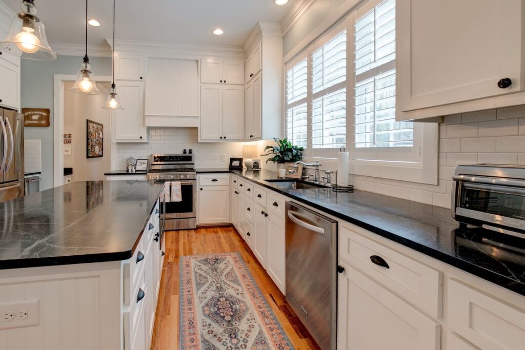 Modern white kitchen with stainless steel appliances, dark countertops, pendant lighting, and large windows with shutters.