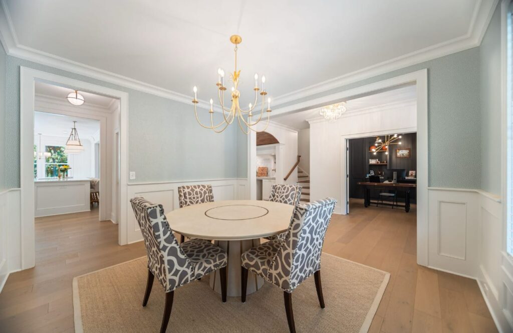 Elegant dining room with round table, patterned chairs, gold chandelier, and light blue walls with white wainscoting.