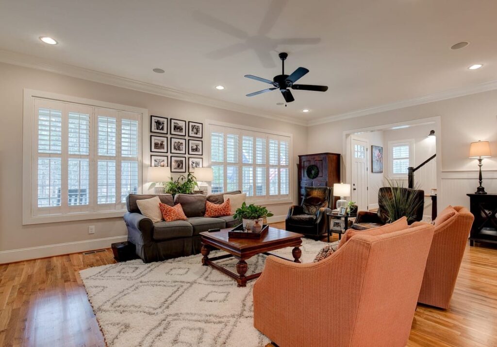 Bright living room with gray sofa, orange chairs, wooden coffee table, and large shuttered windows overlooking water.