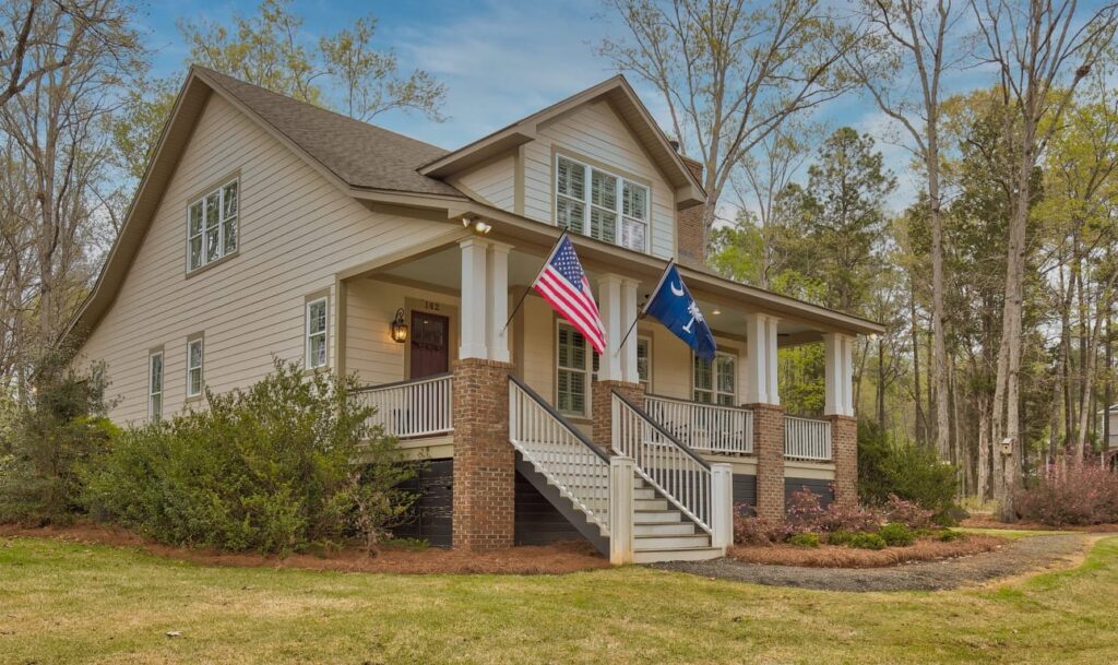Two-story white farmhouse with brick foundation, front porch, American and blue flags, surrounded by bare trees.