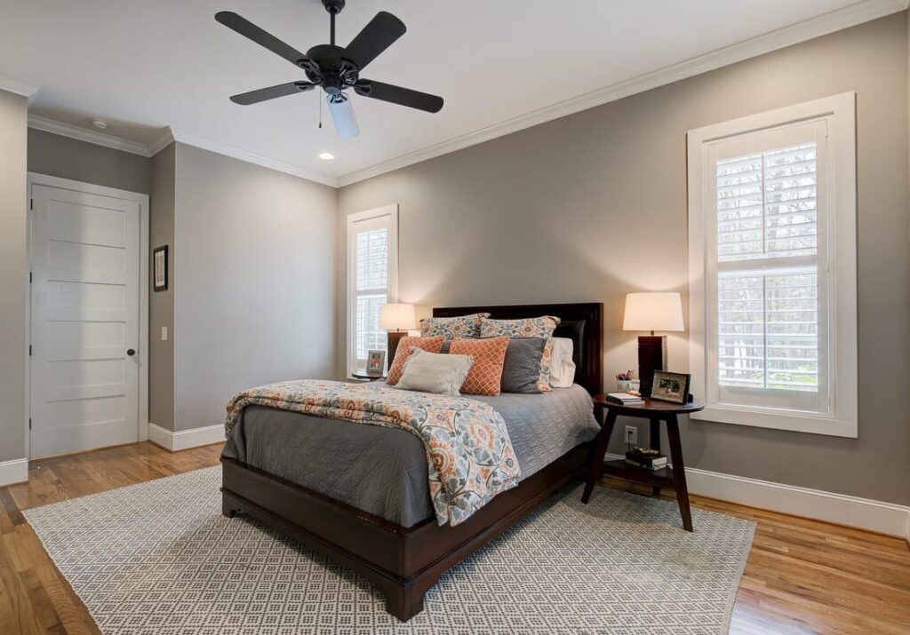 Modern bedroom with dark wood bed, gray bedding, orange patterned pillows, ceiling fan, nightstands with lamps, and large windows.