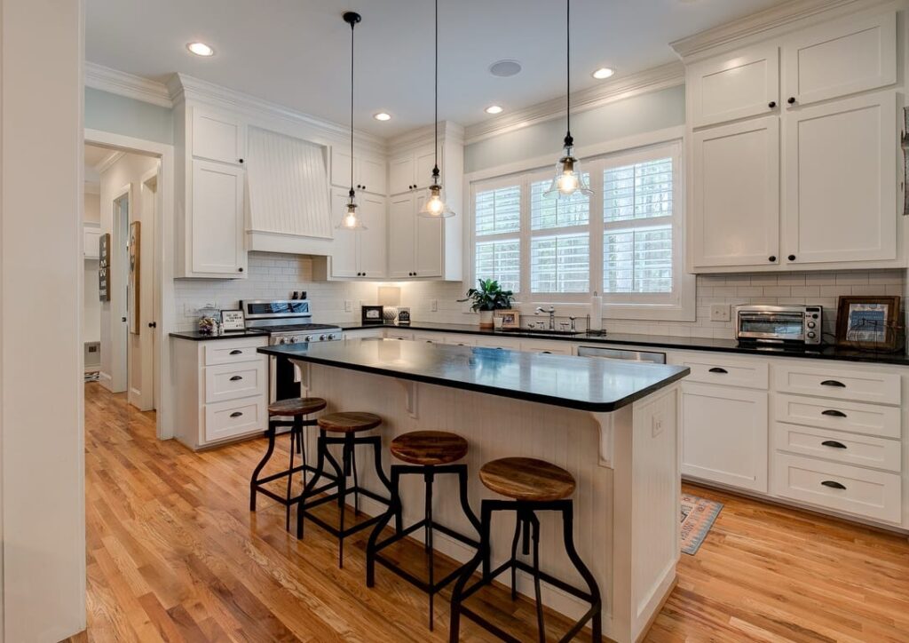 Modern white kitchen with black countertops, island seating, pendant lights, and hardwood floors.