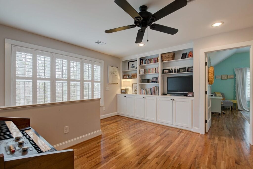 Modern living room with white built-in shelving, TV, wooden floors, piano, ceiling fan, and shuttered windows.