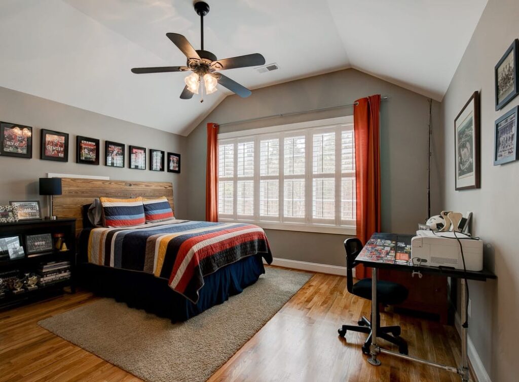 Modern bedroom with striped bedding, wooden desk, ceiling fan, and large shuttered window with orange curtains.