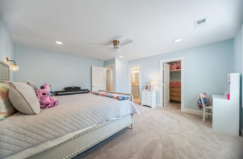 A spacious child's bedroom with light blue walls, a queen bed with gray bedding, pink stuffed animals, white furniture, and carpet flooring.