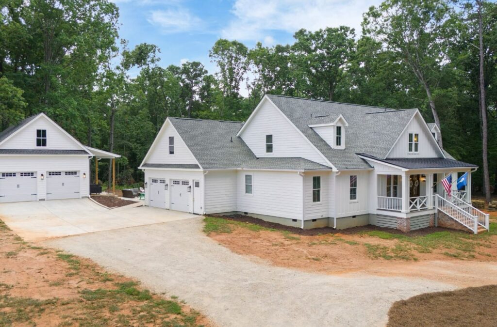 White farmhouse with front porch, wraparound deck, and detached two-car garage on tree-lined lot.
