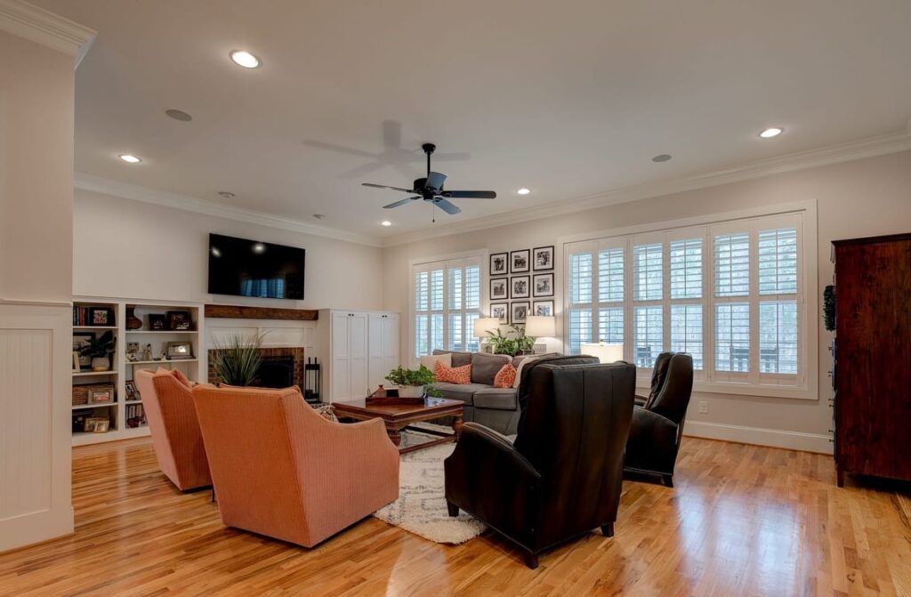 Modern living room with orange and black armchairs, fireplace, built-in shelving, ceiling fan, and large shuttered windows.