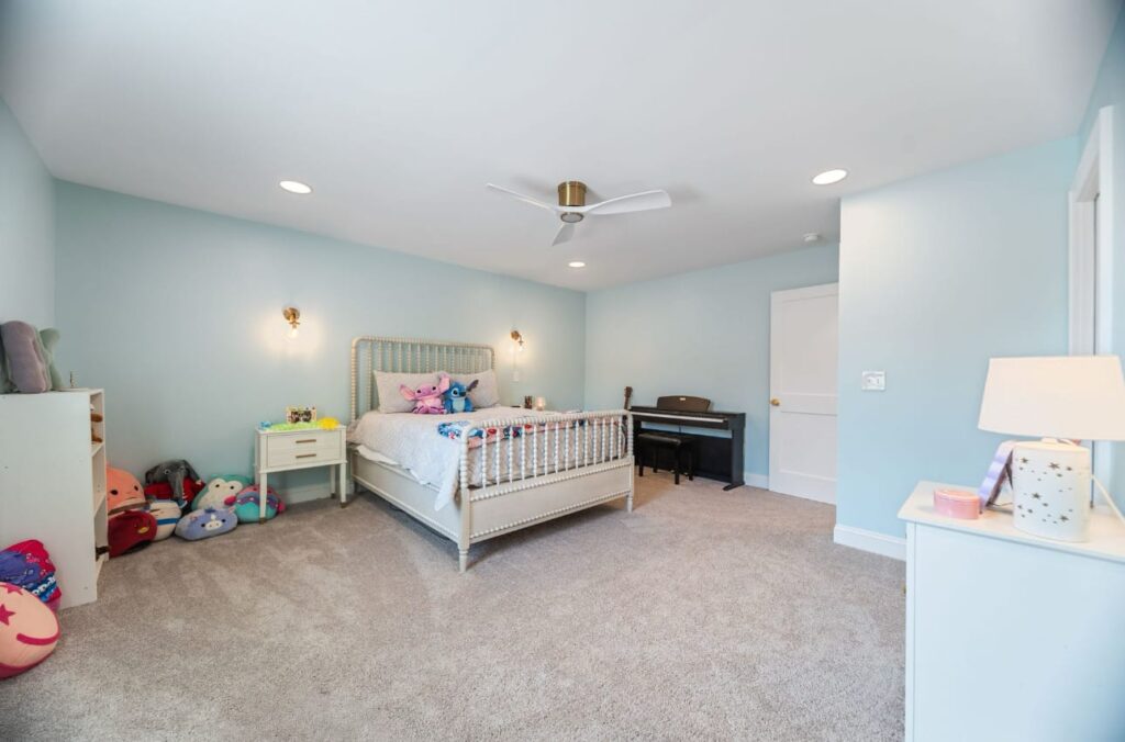 Spacious child's bedroom with light blue walls, white bed, carpet flooring, ceiling fan, and various toys and furniture.