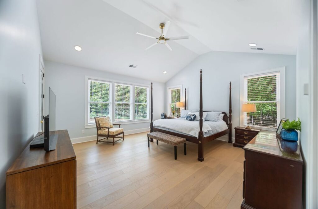 Spacious master bedroom with four-poster bed, hardwood floors, ceiling fan, and multiple windows overlooking greenery.