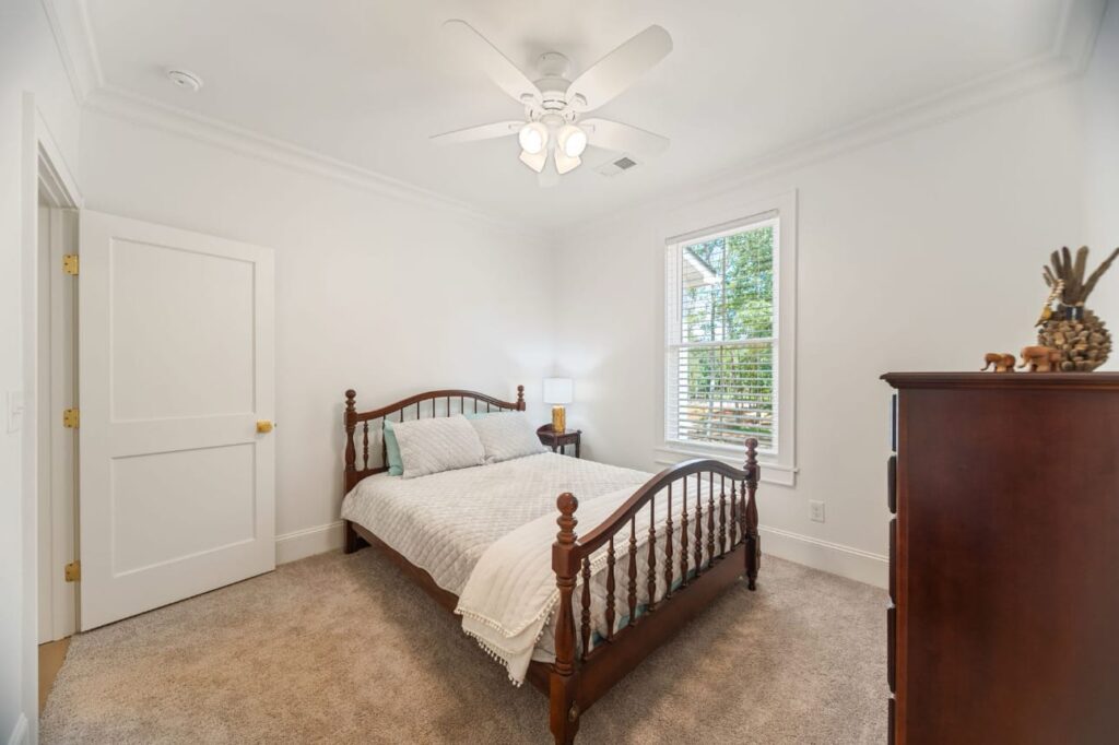 Bright bedroom with wooden bed frame, white bedding, beige carpet, dark wood dresser, ceiling fan, and large window.