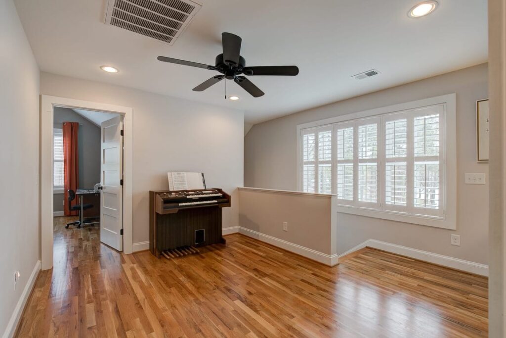 Spacious room with hardwood floors, black ceiling fan, electronic organ, white shutters, and open doorway to adjacent office space.
