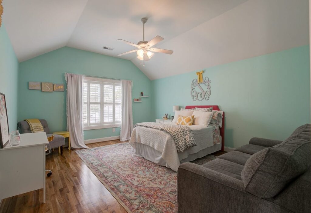 A bright turquoise bedroom with vaulted ceilings, featuring a bed with red headboard, desk area, seating nook by bay window, and wooden flooring.