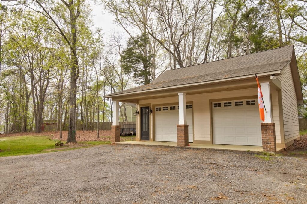 Modern single-story garage with brick foundation, white siding, and two white garage doors, surrounded by bare trees on a gravel driveway.