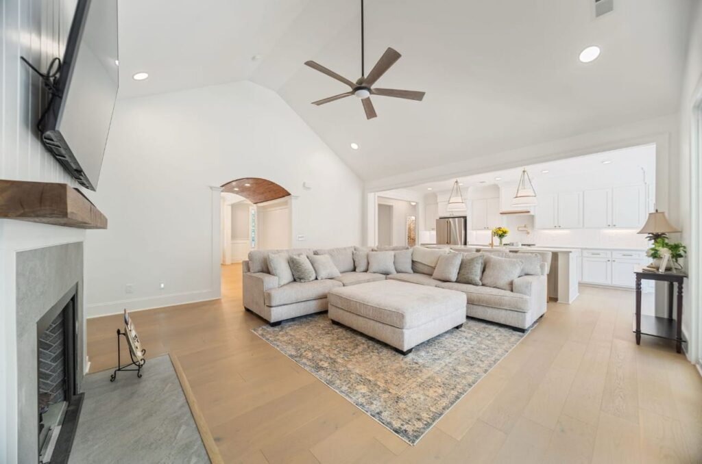Modern open-concept living room with sectional sofa, fireplace, ceiling fan, and kitchen visible in background.