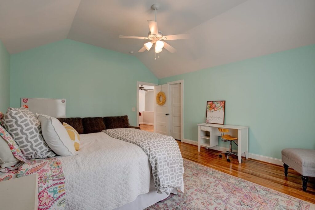 A bright turquoise bedroom with a bed, white desk, ceiling fan, and hardwood flooring.