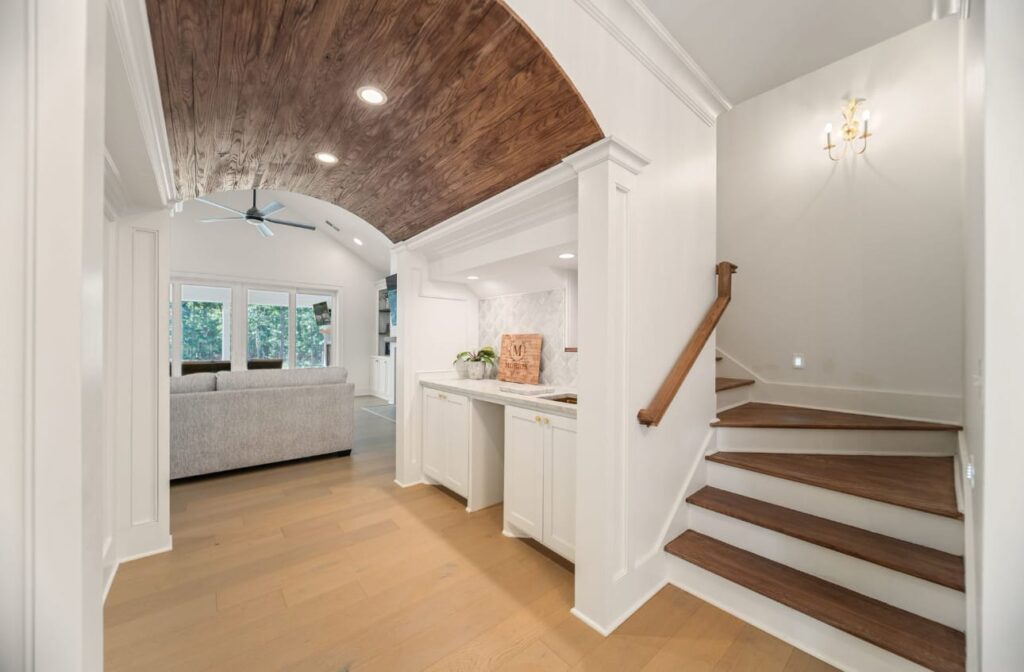 Modern home entryway with curved wood ceiling, white cabinetry, wooden staircase, and open living area with windows.
