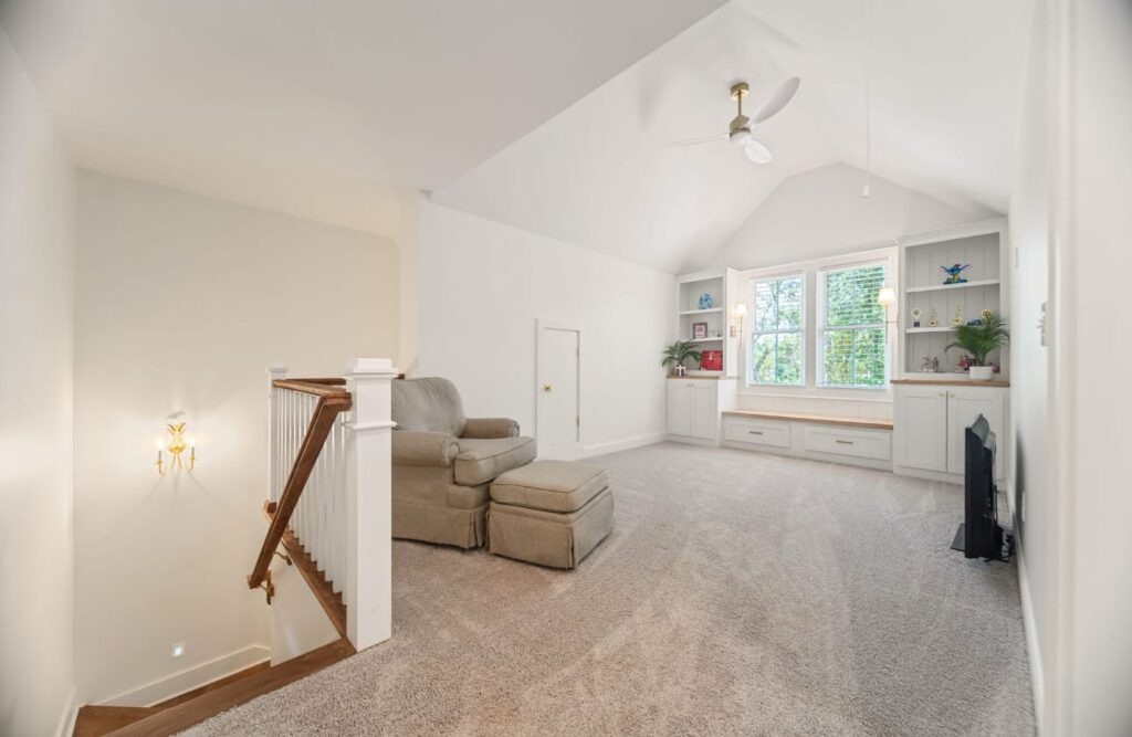 Bright attic bedroom with vaulted ceiling, built-in shelving, armchair, and natural light from windows.