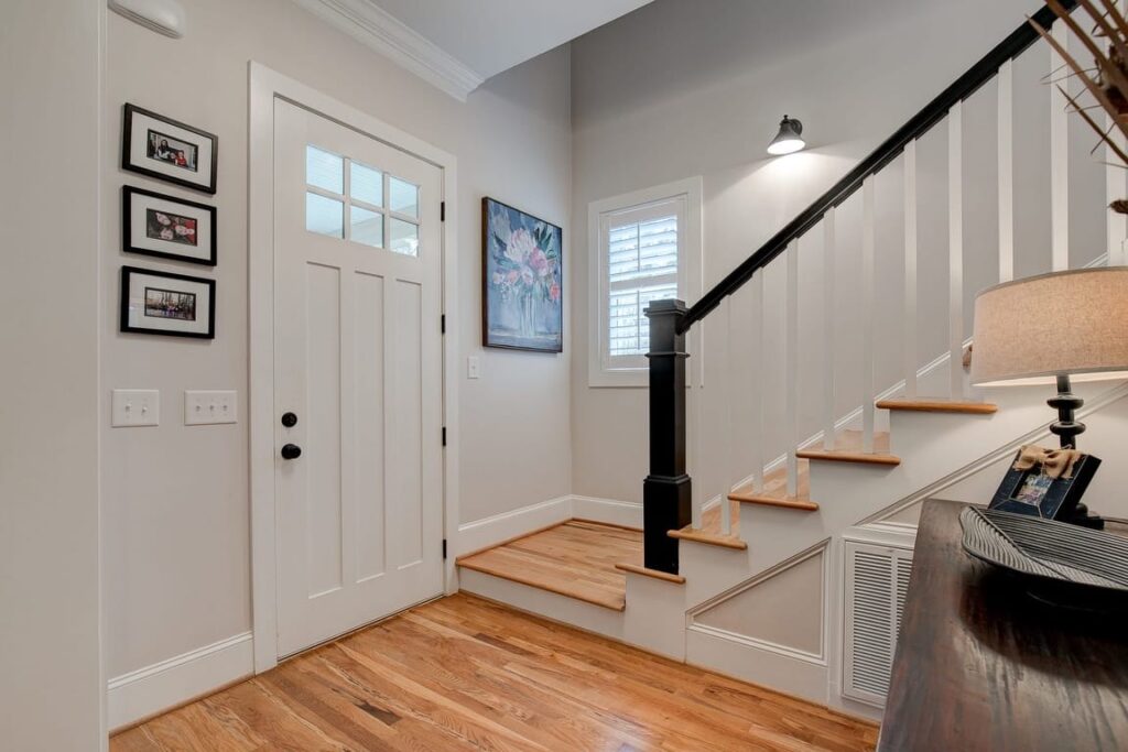 Modern foyer with wooden staircase, white walls, framed photos, front door with glass panels, and black stair railing.
