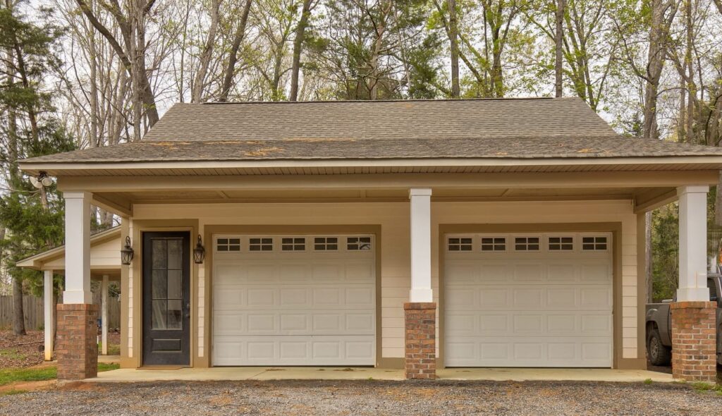 Two-car garage with white doors, brick pillars, and covered portico surrounded by bare trees.