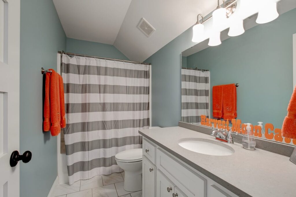 Modern bathroom with turquoise walls, white vanity, dual sinks, striped shower curtain, and orange towel accents.