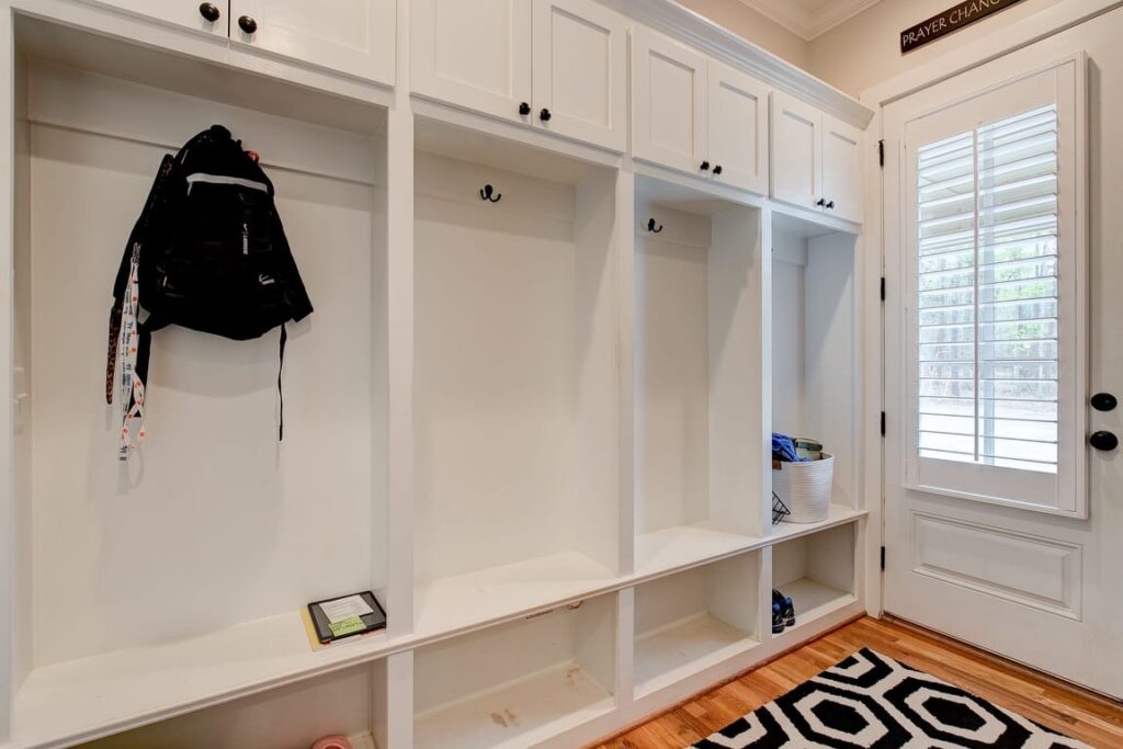 Modern mudroom with white built-in cubbies, wooden shelving, black backpack, and shuttered door leading outside.