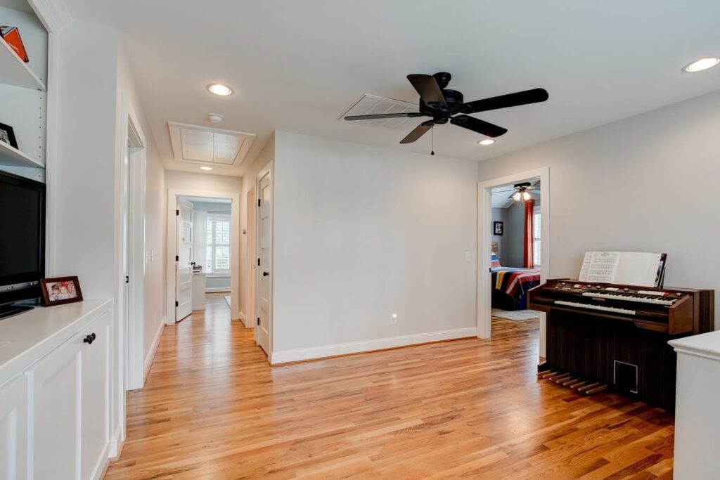 Spacious home music room with wooden floors, ceiling fan, organ keyboard, TV, and hallway leading to other rooms.