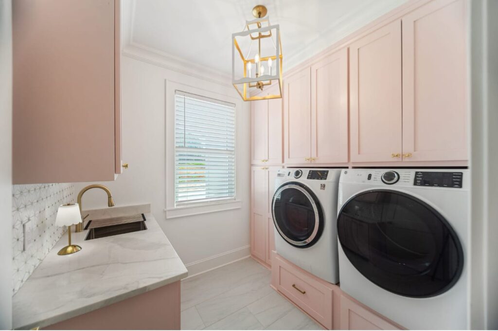 Modern laundry room with pink cabinetry, white marble countertops, stainless steel washer and dryer, and brass fixtures.