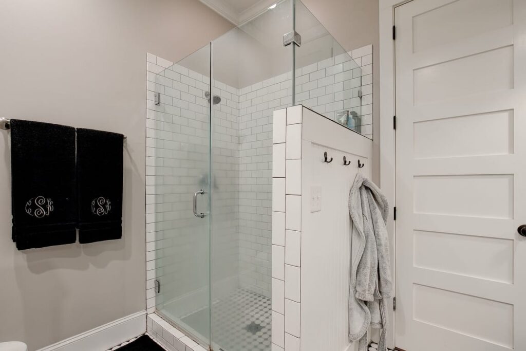 Modern bathroom with frameless glass shower enclosure, white subway tile, black monogrammed towels, and white shaker cabinet door.