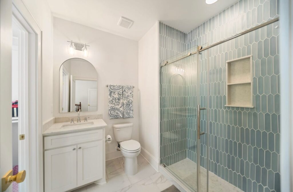 Modern bathroom with white vanity, toilet, and walk-in shower with green geometric tile and glass enclosure.