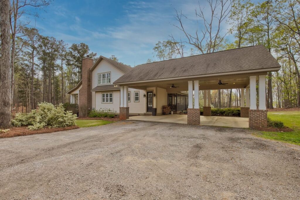 Modern farmhouse with brick and white siding, covered porch, and gravel driveway surrounded by tall trees.